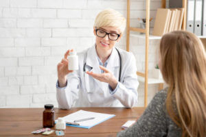 Pharmaceuticals, prescription and doctor recommendation. Friendly adult woman therapist in glasses and white coat shows jar of medicines to patient in interior of office in clinic, free space